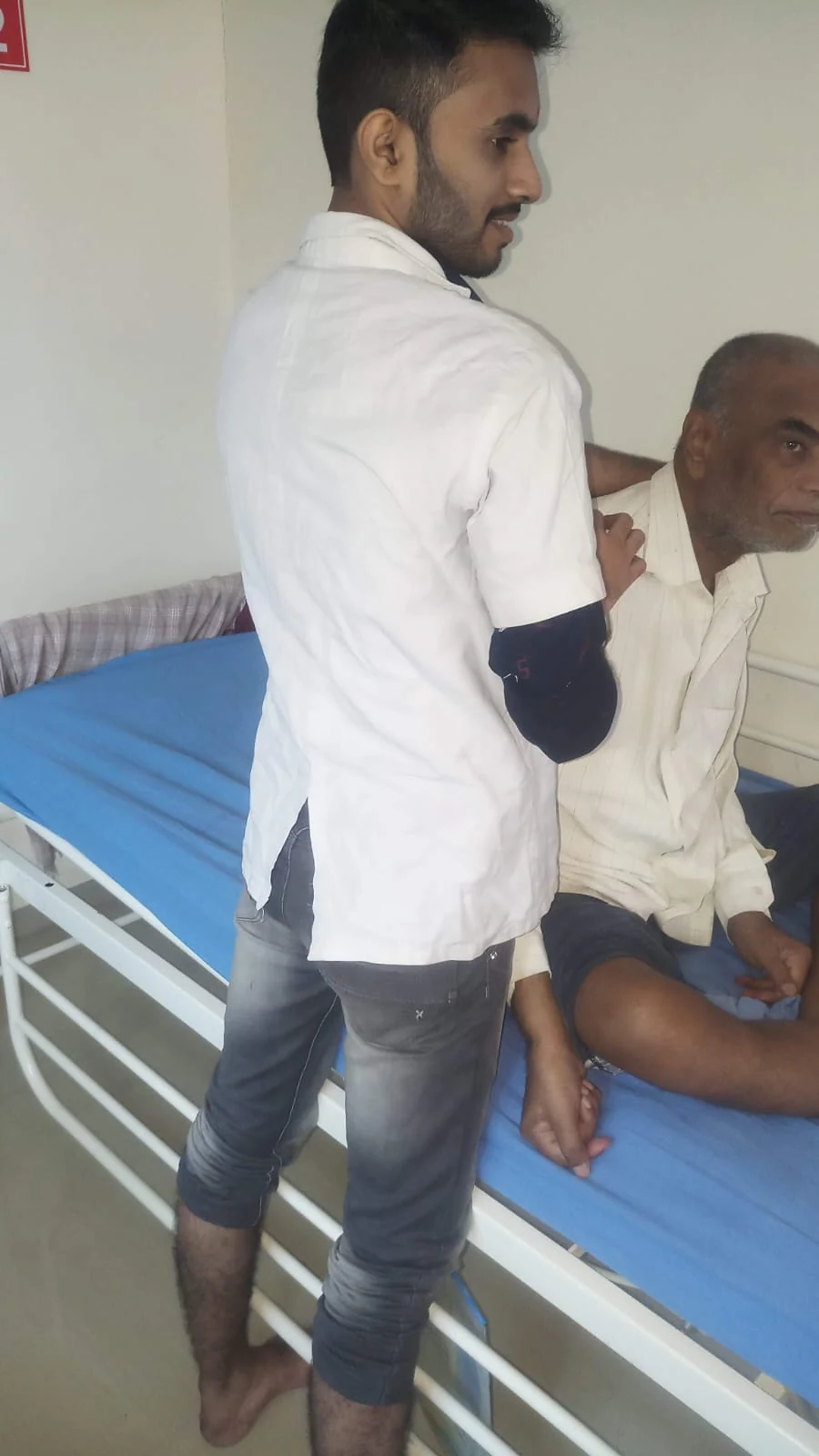 A care worker supporting a seated man at the bedside at Jeevan Jyoti
